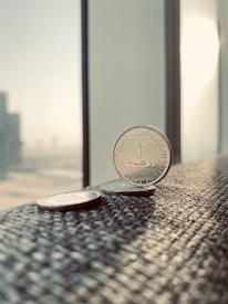 Several coins are placed on a textured surface near a window, capturing a bright, sunlit environment. The primary focus is on a coin from the United Arab Emirates, displaying Arabic script. The background features a blurred cityscape with tall buildings visible through the window.