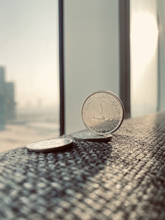 Several coins are placed on a textured surface near a window, capturing a bright, sunlit environment. The primary focus is on a coin from the United Arab Emirates, displaying Arabic script. The background features a blurred cityscape with tall buildings visible through the window.