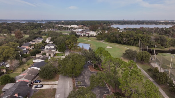 An aerial view of a suburban neighborhood next to a golf course. The landscape includes numerous houses surrounded by lush vegetation and ample greenery. Beyond the residential area, there is a well-maintained golf course with visible sand traps and a small water feature. In the distance, a body of water is seen, lined with trees on its shore.