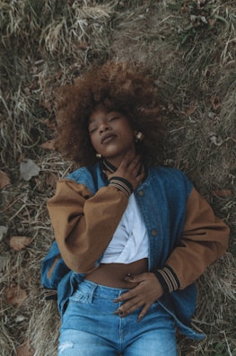 A person lies on their back on a bed of dry grass, wearing a denim and brown jacket over a white shirt and blue jeans. Their left hand rests on their stomach while the right hand lightly touches their neck. The person's eyes are closed and they appear calm and relaxed.