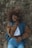 A person lies on their back on a bed of dry grass, wearing a denim and brown jacket over a white shirt and blue jeans. Their left hand rests on their stomach while the right hand lightly touches their neck. The person's eyes are closed and they appear calm and relaxed.