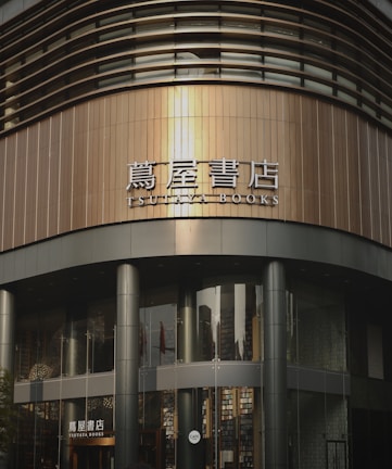 A modern bookstore with a curved, sleek architectural design featuring large glass windows and metal panels. The signage displays both Japanese characters and English text that reads 'TSUTAYA BOOKS'.