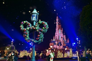 A festive night scene featuring a castle illuminated with blue and pink lights, surrounded by decorated Christmas trees. In the foreground, a lamppost is adorned with a wreath in the shape of Mickey Mouse's head, made of greenery and colorful ornaments. The sky is dark with snowflakes gently falling, and people are walking and enjoying the atmosphere.