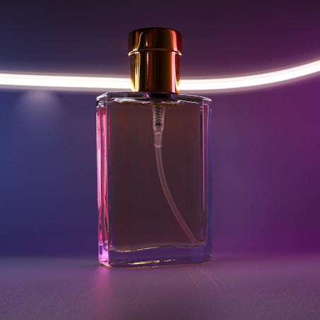 A transparent glass perfume bottle with a gold cap, highlighted by a soft, pink and purple ambient light. The backdrop features a neon-like horizontal light streak, adding a modern and stylish touch.