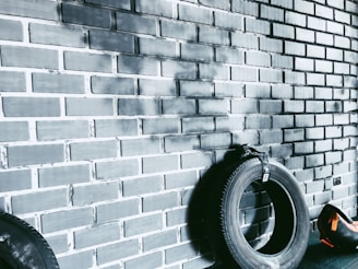 A dark tire is leaning against a painted brick wall with a black and gray gradient. Another tire is partially visible on the left. A small orange and black sports bag is situated on the floor to the right, near the tire.