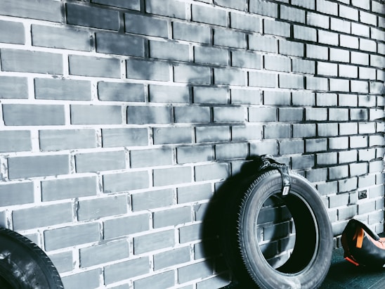 A dark tire is leaning against a painted brick wall with a black and gray gradient. Another tire is partially visible on the left. A small orange and black sports bag is situated on the floor to the right, near the tire.