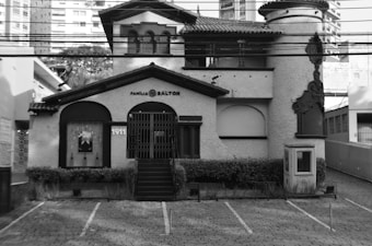 A historic, ornate building with 'Família Salton' signage, featuring classic architectural details like arched windows and decorative borders. The structure has a textured façade, dark accents, and multiple levels including a tower on the right. The outdoor area includes a small landscaped garden and a cobblestone driveway.
