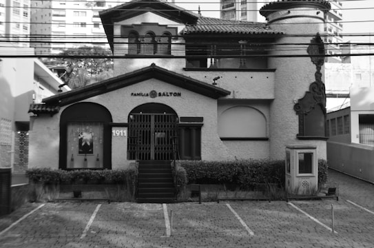 A historic, ornate building with 'Fam&iacute;lia Salton' signage, featuring classic architectural details like arched windows and decorative borders. The structure has a textured fa&ccedil;ade, dark accents, and multiple levels including a tower on the right. The outdoor area includes a small landscaped garden and a cobblestone driveway.