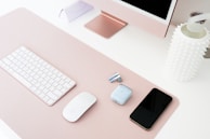 A neatly organized workspace featuring a pink desk setup. A white wireless keyboard and mouse are arranged neatly alongside a smartphone, earphones, and a charging case. The setup also includes a pink computer monitor and a decorative white vase with a spiky texture.
