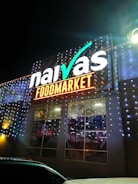 A brightly illuminated storefront sign for a food market is visible. The sign features the brand name in large, white letters with decorative lighting surrounding it. The building is adorned with strings of small, glowing lights, creating a festive appearance at night.