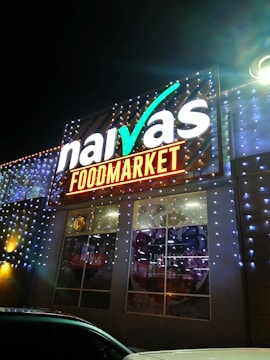 A brightly illuminated storefront sign for a food market is visible. The sign features the brand name in large, white letters with decorative lighting surrounding it. The building is adorned with strings of small, glowing lights, creating a festive appearance at night.