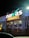 A brightly illuminated storefront sign for a food market is visible. The sign features the brand name in large, white letters with decorative lighting surrounding it. The building is adorned with strings of small, glowing lights, creating a festive appearance at night.