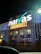 A brightly illuminated storefront sign for a food market is visible. The sign features the brand name in large, white letters with decorative lighting surrounding it. The building is adorned with strings of small, glowing lights, creating a festive appearance at night.