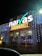 A brightly illuminated storefront sign for a food market is visible. The sign features the brand name in large, white letters with decorative lighting surrounding it. The building is adorned with strings of small, glowing lights, creating a festive appearance at night.