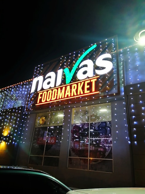 A brightly illuminated storefront sign for a food market is visible. The sign features the brand name in large, white letters with decorative lighting surrounding it. The building is adorned with strings of small, glowing lights, creating a festive appearance at night.