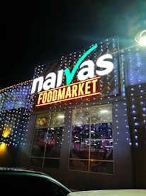 A brightly illuminated storefront sign for a food market is visible. The sign features the brand name in large, white letters with decorative lighting surrounding it. The building is adorned with strings of small, glowing lights, creating a festive appearance at night.