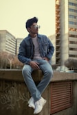 A stylish individual is seated on a concrete ledge, wearing a denim jacket, jeans, and white sneakers. The person sports sunglasses and has neatly groomed hair. The background includes urban high-rise buildings and some graffiti on the wall.