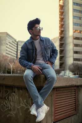 A stylish individual is seated on a concrete ledge, wearing a denim jacket, jeans, and white sneakers. The person sports sunglasses and has neatly groomed hair. The background includes urban high-rise buildings and some graffiti on the wall.