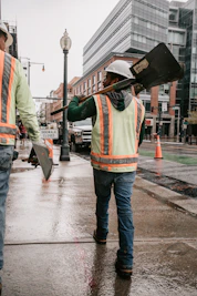 a construction worker carrying a large shovel across a wet street