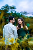 A couple is standing in a natural setting, with the woman lovingly looking at the man, while he is turned towards her. They are surrounded by blurred greenery and have a joyful demeanor.