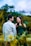 A couple is standing in a natural setting, with the woman lovingly looking at the man, while he is turned towards her. They are surrounded by blurred greenery and have a joyful demeanor.