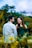 A couple is standing in a natural setting, with the woman lovingly looking at the man, while he is turned towards her. They are surrounded by blurred greenery and have a joyful demeanor.