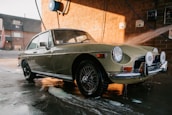 A classic car is being washed at a car wash station. The vehicle has a retro design with circular headlights and chrome details. Water is being sprayed on it, and soap suds can be seen on the pavement. The background features a brick wall and some equipment related to the car wash.