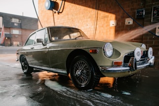 A classic car is being washed at a car wash station. The vehicle has a retro design with circular headlights and chrome details. Water is being sprayed on it, and soap suds can be seen on the pavement. The background features a brick wall and some equipment related to the car wash.
