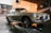 A classic car is being washed at a car wash station. The vehicle has a retro design with circular headlights and chrome details. Water is being sprayed on it, and soap suds can be seen on the pavement. The background features a brick wall and some equipment related to the car wash.