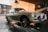 A classic car is being washed at a car wash station. The vehicle has a retro design with circular headlights and chrome details. Water is being sprayed on it, and soap suds can be seen on the pavement. The background features a brick wall and some equipment related to the car wash.