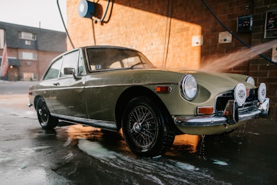 A classic car is being washed at a car wash station. The vehicle has a retro design with circular headlights and chrome details. Water is being sprayed on it, and soap suds can be seen on the pavement. The background features a brick wall and some equipment related to the car wash.