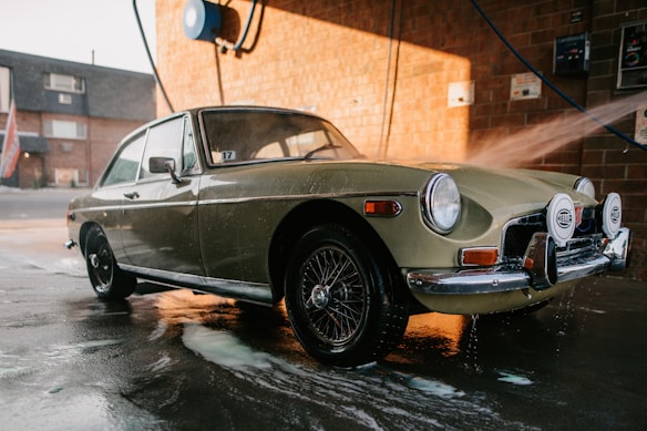 A classic car is being washed at a car wash station. The vehicle has a retro design with circular headlights and chrome details. Water is being sprayed on it, and soap suds can be seen on the pavement. The background features a brick wall and some equipment related to the car wash.