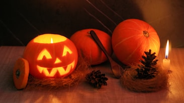 A carved pumpkin with a glowing face is illuminated by a candle inside, creating a warm and festive atmosphere. Two uncarved pumpkins rest nearby, along with a spoon, pine cones, and a small lit candle on a rustic wooden surface. The background is dark, enhancing the soft, flickering light from the candles.
