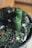 Two green candles are placed upright on a decorative wreath made from evergreen branches, pinecones, and small star-shaped ornaments. The setup is arranged on a metallic tray with intricate patterns. The overall composition evokes a festive and natural aesthetic.
