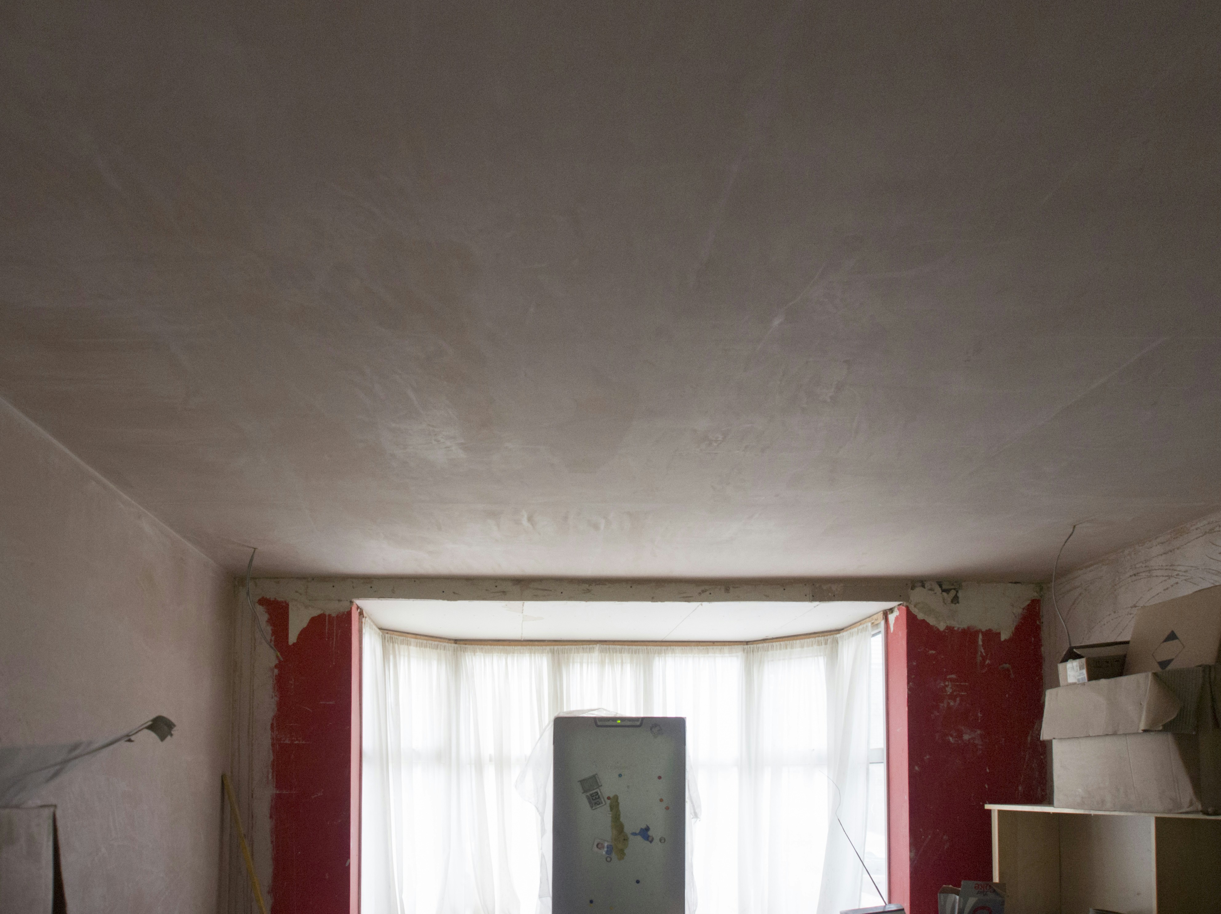 A dimly lit room under renovation, featuring unfinished plaster walls and a large central window with sheer curtains. A refrigerator stands in front of the window, surrounded by various items, including shelving and stacked boxes. Exposed plaster and red paint remnants are visible around the windows.