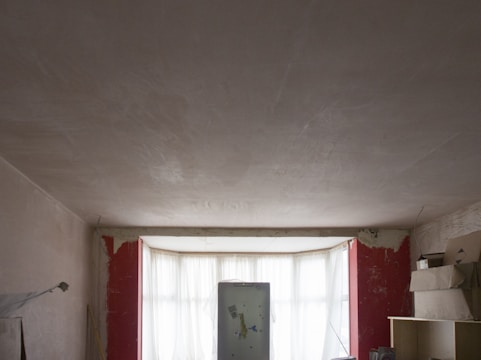 A dimly lit room under renovation, featuring unfinished plaster walls and a large central window with sheer curtains. A refrigerator stands in front of the window, surrounded by various items, including shelving and stacked boxes. Exposed plaster and red paint remnants are visible around the windows.