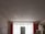 A dimly lit room under renovation, featuring unfinished plaster walls and a large central window with sheer curtains. A refrigerator stands in front of the window, surrounded by various items, including shelving and stacked boxes. Exposed plaster and red paint remnants are visible around the windows.
