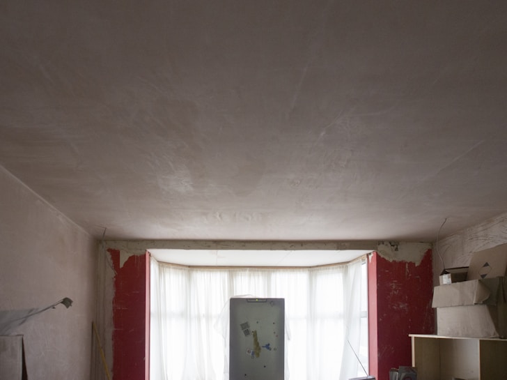 A dimly lit room under renovation, featuring unfinished plaster walls and a large central window with sheer curtains. A refrigerator stands in front of the window, surrounded by various items, including shelving and stacked boxes. Exposed plaster and red paint remnants are visible around the windows.