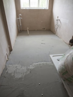 A partially renovated room with freshly laid concrete flooring. The room has bare plastered walls and two exposed pipes for plumbing. A window at the far end allows natural light in, partially illuminating the room. There is building material placed in the corner.
