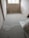 A partially renovated room with freshly laid concrete flooring. The room has bare plastered walls and two exposed pipes for plumbing. A window at the far end allows natural light in, partially illuminating the room. There is building material placed in the corner.