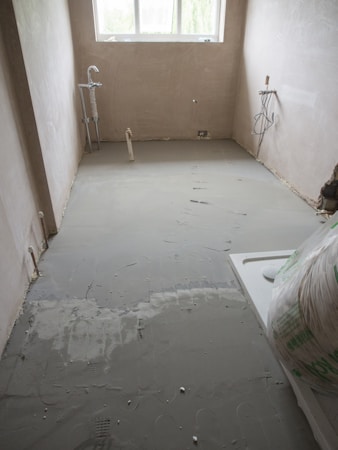 A partially renovated room with freshly laid concrete flooring. The room has bare plastered walls and two exposed pipes for plumbing. A window at the far end allows natural light in, partially illuminating the room. There is building material placed in the corner.