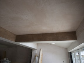 A room under construction with unfinished walls and ceiling. A large wooden beam is visible, spanning across the ceiling. Light enters through a window on the right, partially covered by a plastic sheet. Various construction materials and tools are scattered around.