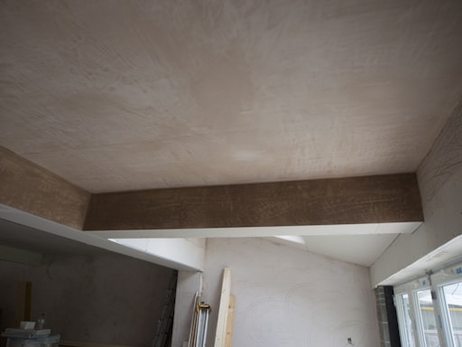 A room under construction with unfinished walls and ceiling. A large wooden beam is visible, spanning across the ceiling. Light enters through a window on the right, partially covered by a plastic sheet. Various construction materials and tools are scattered around.