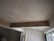 A room under construction with unfinished walls and ceiling. A large wooden beam is visible, spanning across the ceiling. Light enters through a window on the right, partially covered by a plastic sheet. Various construction materials and tools are scattered around.