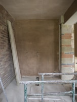 An unfinished room with exposed brick walls, a partially plastered wall, and construction materials scattered on the floor. A green metal framework is in the foreground, possibly scaffolding or a support structure. Electrical wires protrude from the unpainted wall.