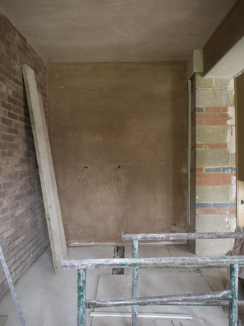 An unfinished room with exposed brick walls, a partially plastered wall, and construction materials scattered on the floor. A green metal framework is in the foreground, possibly scaffolding or a support structure. Electrical wires protrude from the unpainted wall.