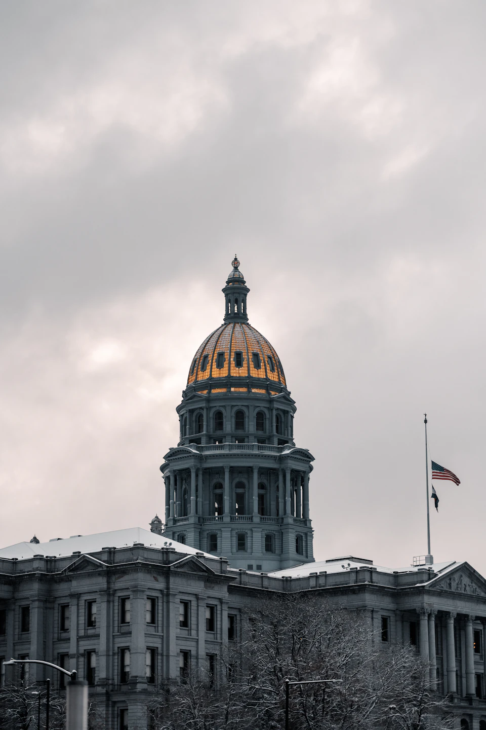 The image depicts a majestic, large building with a golden dome and a flagpole on top. The building is constructed from gray stone and features multiple levels, giving it an imposing presence against the backdrop of a cloudy sky. A street lamp can be seen in front of the building, adding depth to the scene.