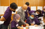A group of people wearing face masks and purple shirts are gathered around a table, engaged in a discussion or collaborative activity. They appear to be in a learning or workshop setting, with papers and bottles on the table.