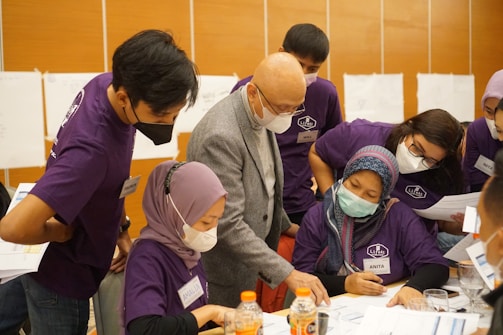 A group of people wearing face masks and purple shirts are gathered around a table, engaged in a discussion or collaborative activity. They appear to be in a learning or workshop setting, with papers and bottles on the table.