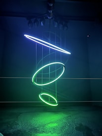 A dark room features an abstract, suspended installation with three glowing neon rings, each in a vivid green hue. A diagonal neon tube in blue hovers above, creating a modern, minimalist effect. The subtle reflections on the floor enhance the futuristic atmosphere.